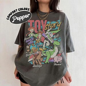May include: A charcoal gray short-sleeve t-shirt featuring a graphic print of Toy Story characters including Jessie, Woody, Buzz Lightyear, Slinky Dog, Bullseye, Rex, and Hamm. The text reads Comfort Colors Pepper, Toy Story, Jessie Since 1995, and names.