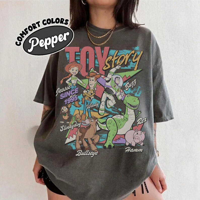 May include: A charcoal gray short-sleeve t-shirt featuring a graphic print of Toy Story characters including Jessie, Woody, Buzz Lightyear, Slinky Dog, Bullseye, Rex, and Hamm. The text reads Comfort Colors Pepper, Toy Story, Jessie Since 1995, and names.