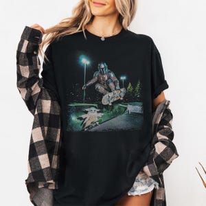 Op de afbeelding: Zwart t-shirt met een afbeelding van The Mandalorian en Grogu die 's nachts in een skatepark skaten. The Mandalorian is in de lucht op een skateboard, terwijl Grogu eronder skatet. De achtergrond bevat bomen en straatverlichting.
