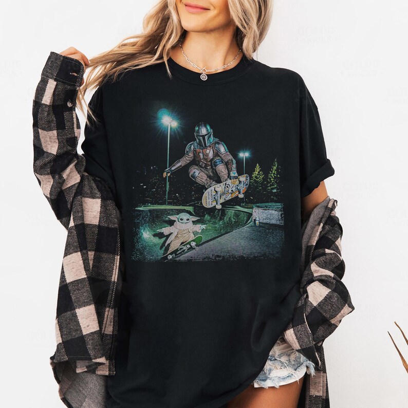 Op de afbeelding: Zwart t-shirt met een afbeelding van The Mandalorian en Grogu die 's nachts in een skatepark skaten. The Mandalorian is in de lucht op een skateboard, terwijl Grogu eronder skatet. De achtergrond bevat bomen en straatverlichting.