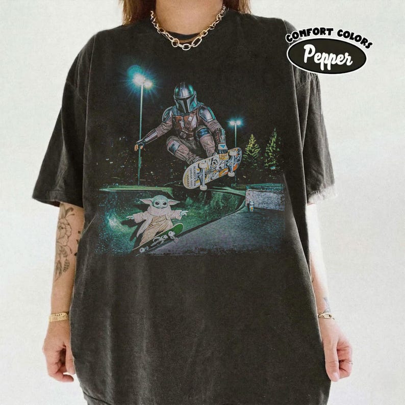 Op de afbeelding: Donkergrijs t-shirt met een afbeelding van The Mandalorian en Grogu die skaten in een skatepark. Het ontwerp bevat een nachtsc&egrave;ne met groene en blauwe accenten. De tekst "COMFORT COLORS Pepper" staat rechtsboven.