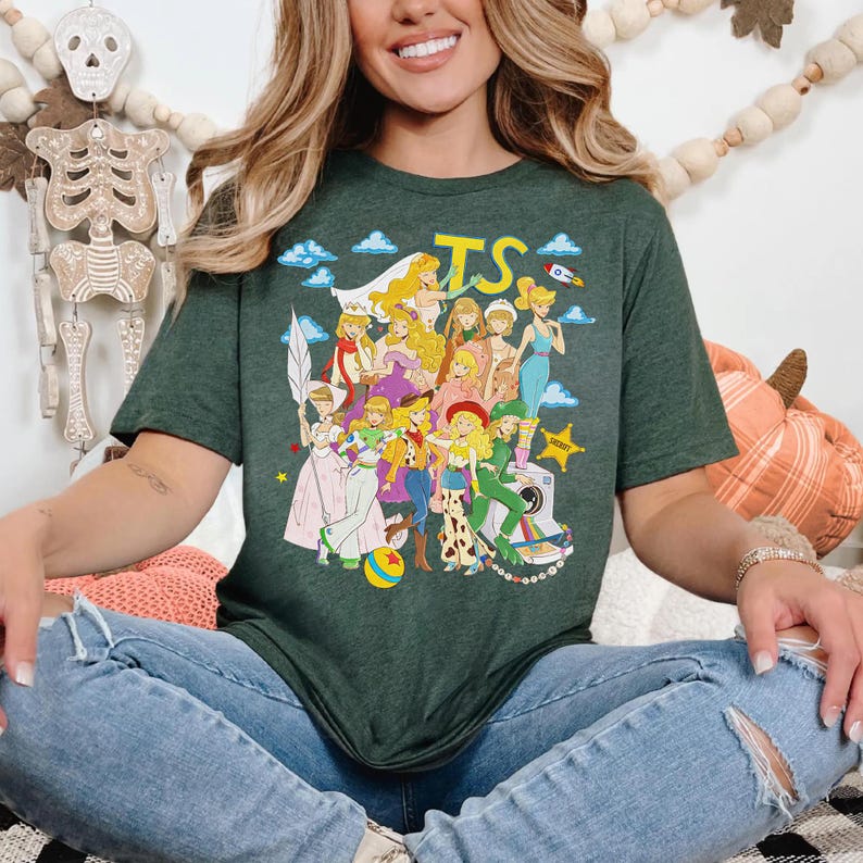 Puede incluir: Una camiseta de manga corta de color verde oscuro con un estampado gr&aacute;fico colorido de varios personajes animados con diversos atuendos, incluyendo una vaquera, un guardi&aacute;n espacial y un sheriff, con el texto amarillo TS y nubes azules.