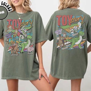 May include: A moss green t-shirt featuring a graphic print of Toy Story characters including Woody, Buzz Lightyear, Jessie, Rex, Bullseye, Slinky Dog, and Hamm. The design includes the text Toy Story and Since 1995 in a colorful, retro-inspired font style.