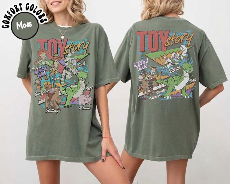 May include: A moss green t-shirt featuring a graphic print of Toy Story characters including Woody, Buzz Lightyear, Jessie, Rex, Bullseye, Slinky Dog, and Hamm. The design includes the text Toy Story and Since 1995 in a colorful, retro-inspired font style.