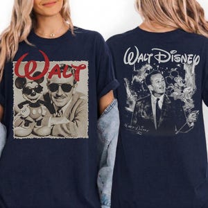 May include: A navy blue t-shirt featuring a front graphic of Walt Disney and Mickey Mouse wearing sunglasses with the red text Walt above them. The back of the shirt displays a black and white illustration of Walt Disney with Mickey Mouse.