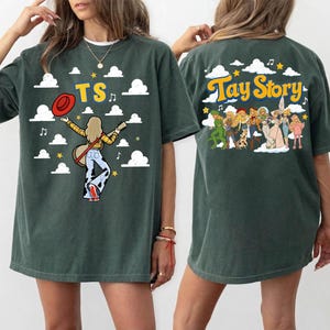 May include: A dark green short-sleeve t-shirt featuring a graphic print. The front displays the text TS with a figure holding a guitar and hat amidst clouds. The back shows the text Tay Story with a group of illustrated figures and clouds.