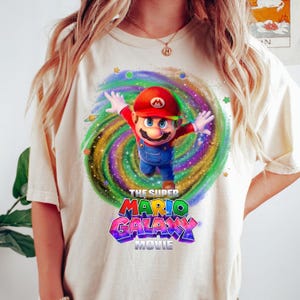 May include: A cream-colored t-shirt featuring a graphic of Mario from the Super Mario Galaxy movie. Mario is depicted with arms outstretched against a swirling, colorful galaxy background with stars and planets. The text reads The Super Mario Galaxy Movie.
