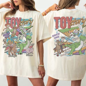 May include: An off-white t-shirt featuring a large graphic print of Toy Story characters including Woody, Buzz Lightyear, Jessie, Rex, Bullseye, Slinky Dog, and Hamm. The design includes the text Toy Story and Since 1995 with colorful geometric shapes.