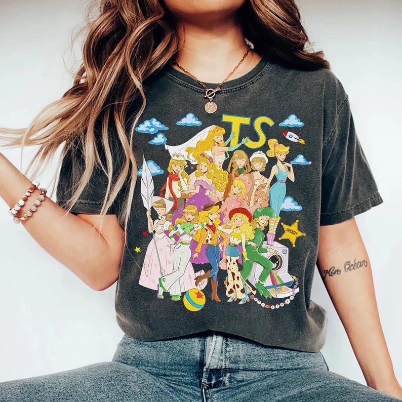 Puede incluir: Una camiseta gris carb&oacute;n de manga corta con un estampado gr&aacute;fico de varios personajes animados con diversos atuendos, incluyendo un traje espacial, ropa de vaquero y un vestido blanco. El texto TS est&aacute; impreso en amarillo sobre ellos.