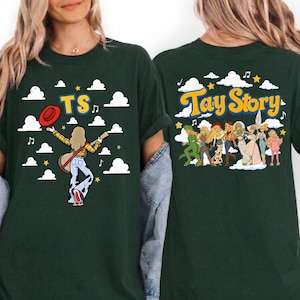May include: A dark green t-shirt featuring a graphic print. The front shows a figure with a guitar, a red hat, and the text TS. The back displays a group of illustrated figures and the text Tay Story, both set against a background of clouds.