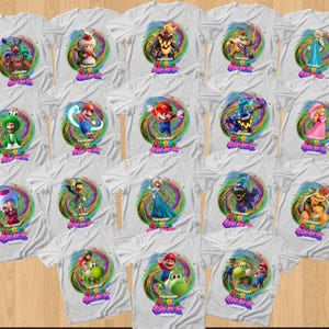 May include: A collection of white t-shirts featuring various Super Mario Galaxy character graphics. Each shirt displays a colorful, swirling cosmic background behind a specific character, with the Super Mario Galaxy logo printed below the artwork.