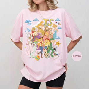 May include: A light pink t-shirt featuring a graphic print of various animated female characters in diverse outfits. The design includes the yellow text TS, blue clouds, a small rocket, a yellow star, and a colorful ball. The shirt has short sleeves.