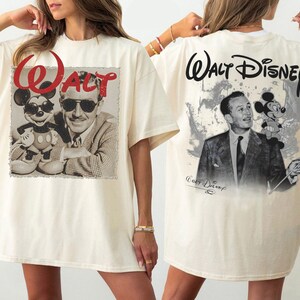May include: An off-white t-shirt featuring a front graphic of Walt Disney and Mickey Mouse with the red text "Walt" above them. The back graphic displays Walt Disney and Mickey Mouse with the black text "Walt Disney" printed above.