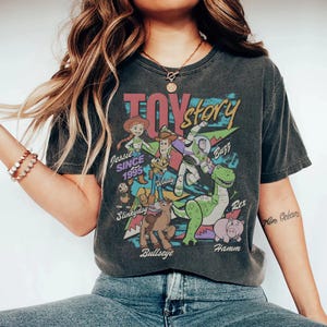 May include: A charcoal gray short-sleeve t-shirt featuring a colorful graphic print of Toy Story characters including Jessie, Woody, Buzz Lightyear, Rex, Hamm, Bullseye, and Slinky Dog. The text reads Toy Story, Jessie SINCE 1995, Woody, Buzz, Rex, Hamm, and Bullseye.