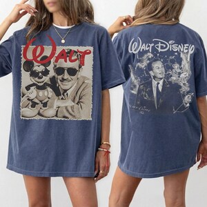 May include: A blue t-shirt featuring a graphic on the front of Walt Disney and Mickey Mouse wearing sunglasses with the red text WALT above them. The back of the t-shirt displays a graphic of Walt Disney with the text WALT DISNEY printed above.