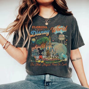 May include: A charcoal gray short-sleeve t-shirt featuring a colorful graphic print of Florida Disney World. The design includes Mickey Mouse, the castle, Epcot, and the text Florida Est. 1971 Disney World The Most Magical Place On Earth.