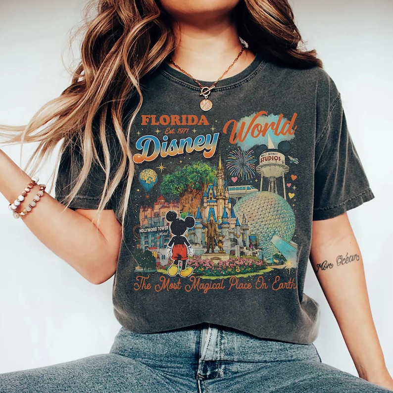 May include: A charcoal gray short-sleeve t-shirt featuring a colorful graphic print of Florida Disney World. The design includes Mickey Mouse, the castle, Epcot, and the text Florida Est. 1971 Disney World The Most Magical Place On Earth.
