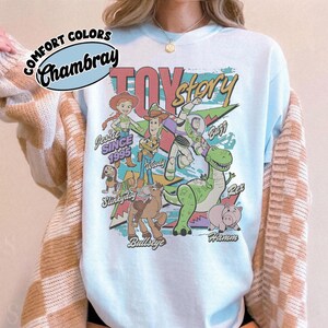 May include: A light blue t-shirt featuring a graphic print of Toy Story characters including Woody, Buzz Lightyear, Jessie, Slinky Dog, Bullseye, Rex, and Hamm. The text reads Toy Story, Jessie Since 1995, Woody, Buzz, Slinky Dog, Bullseye, and Hamm.