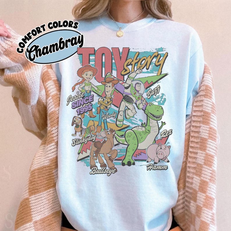 May include: A light blue t-shirt featuring a graphic print of Toy Story characters including Woody, Buzz Lightyear, Jessie, Slinky Dog, Bullseye, Rex, and Hamm. The text reads Toy Story, Jessie Since 1995, Woody, Buzz, Slinky Dog, Bullseye, and Hamm.