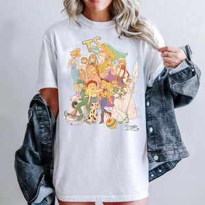 Puede incluir: Una camiseta blanca de manga corta con una colorida ilustraci&oacute;n gr&aacute;fica de varios personajes animados con diversos atuendos, incluyendo un logotipo amarillo 'TS' en la parte superior, una c&aacute;mara vintage y una pelota amarilla y azul.