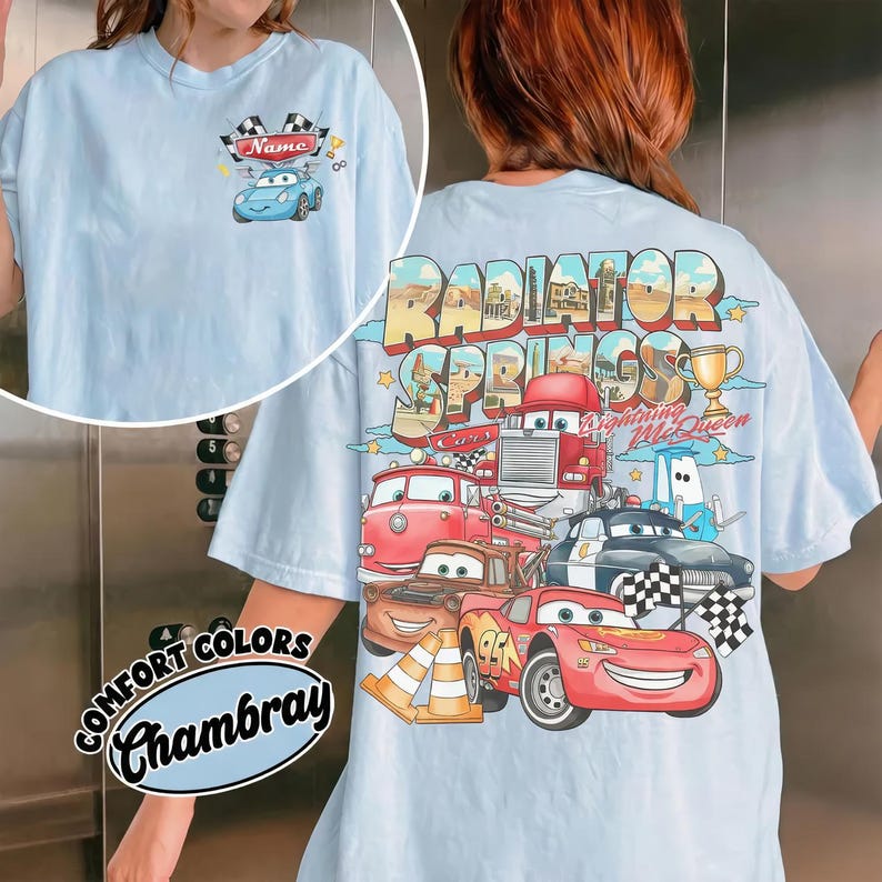 Puede incluir: Una camiseta azul claro con un peque&ntilde;o gr&aacute;fico en el pecho que muestra un coche azul y el texto Name. La parte trasera presenta un gran gr&aacute;fico de Radiator Springs con varios coches de dibujos animados, incluyendo un coche de carreras rojo.