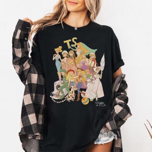 Puede incluir: Una camiseta negra con un estampado gr&aacute;fico de varios personajes femeninos ilustrados con diversos atuendos, las letras TS en amarillo y una pelota colorida. La camiseta se lleva debajo de una camisa de cuadros negros y blancos.