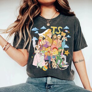 May include: A charcoal gray short-sleeve t-shirt featuring a graphic print of multiple animated characters dressed in various outfits. The design includes the yellow text TS, blue clouds, a rocket, a sheriff star, and a colorful ball at the bottom.