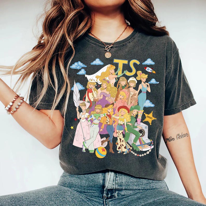 May include: A charcoal gray short-sleeve t-shirt featuring a graphic print of multiple animated characters dressed in various outfits. The design includes the yellow text TS, blue clouds, a rocket, a sheriff star, and a colorful ball at the bottom.