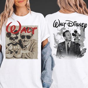 May include: A white t-shirt featuring a graphic print on the front and back. The front displays a black and white image of Walt Disney with Mickey Mouse, both wearing sunglasses, under the red text "WALT". The back shows Walt Disney with Mickey Mouse and the text "Walt Disney".