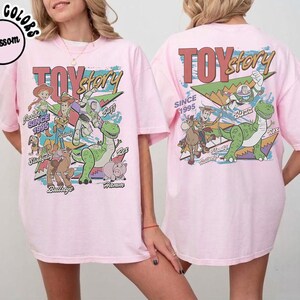 May include: A light pink t-shirt featuring a graphic print of Toy Story characters including Woody, Buzz Lightyear, Jessie, Rex, Slinky Dog, Bullseye, and Hamm. The design includes the text Toy Story and Since 1995 with colorful geometric shapes.