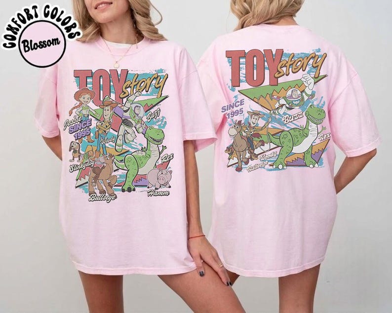 May include: A light pink t-shirt featuring a graphic print of Toy Story characters including Woody, Buzz Lightyear, Jessie, Rex, Slinky Dog, Bullseye, and Hamm. The design includes the text Toy Story and Since 1995 with colorful geometric shapes.