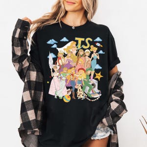May include: A black t-shirt featuring a graphic print of various animated characters dressed in diverse outfits, including a space suit, cowgirl attire, and gowns. The design includes the text TS, a rocket, clouds, a star, and a camera.