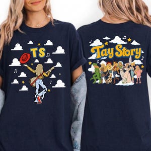 May include: A navy blue t-shirt featuring a graphic print on the front and back. The front shows a figure with a guitar and the text TS. The back displays a group of characters and the text Tay Story, both set against a background of clouds.