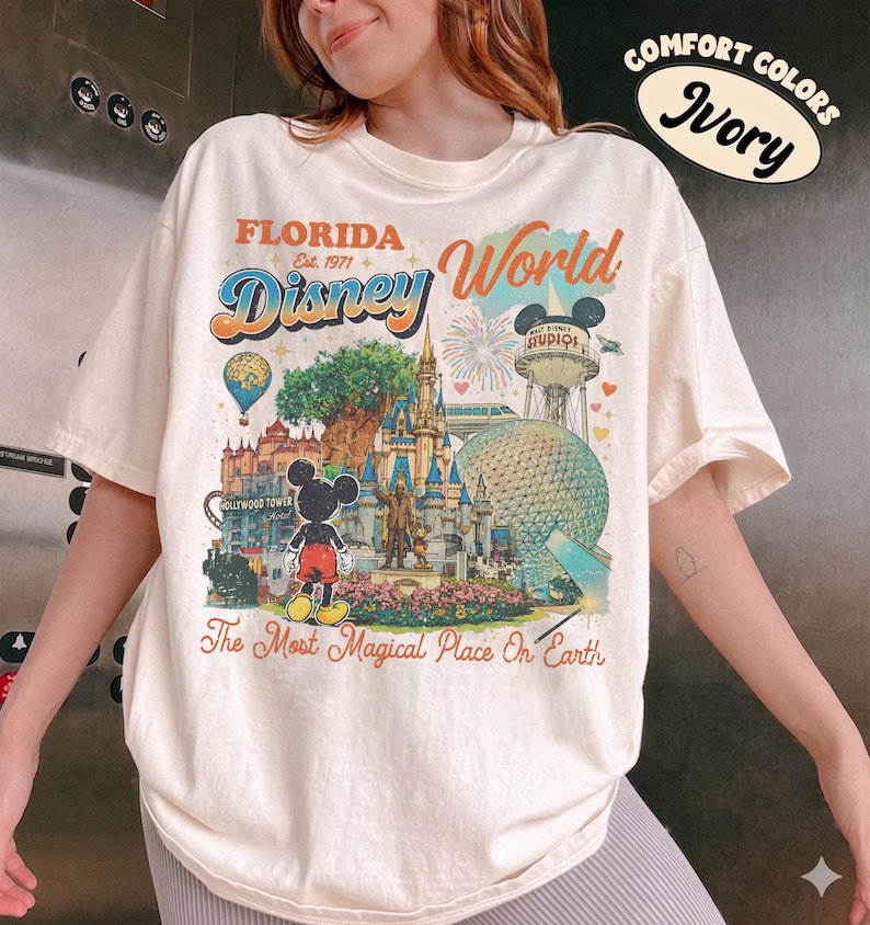 May include: An ivory t-shirt featuring a colorful graphic print of Florida Disney World landmarks, including the castle, Epcot sphere, and Hollywood Tower. The text reads Florida, Disney World, Est. 1971, and The Most Magical Place On Earth.