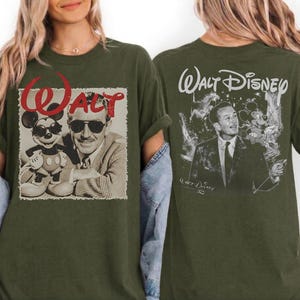 May include: An olive green t-shirt featuring a front graphic of Walt Disney and Mickey Mouse wearing sunglasses with the red text Walt above them, and a back graphic of Walt Disney in a suit with the text Walt Disney printed in white script.