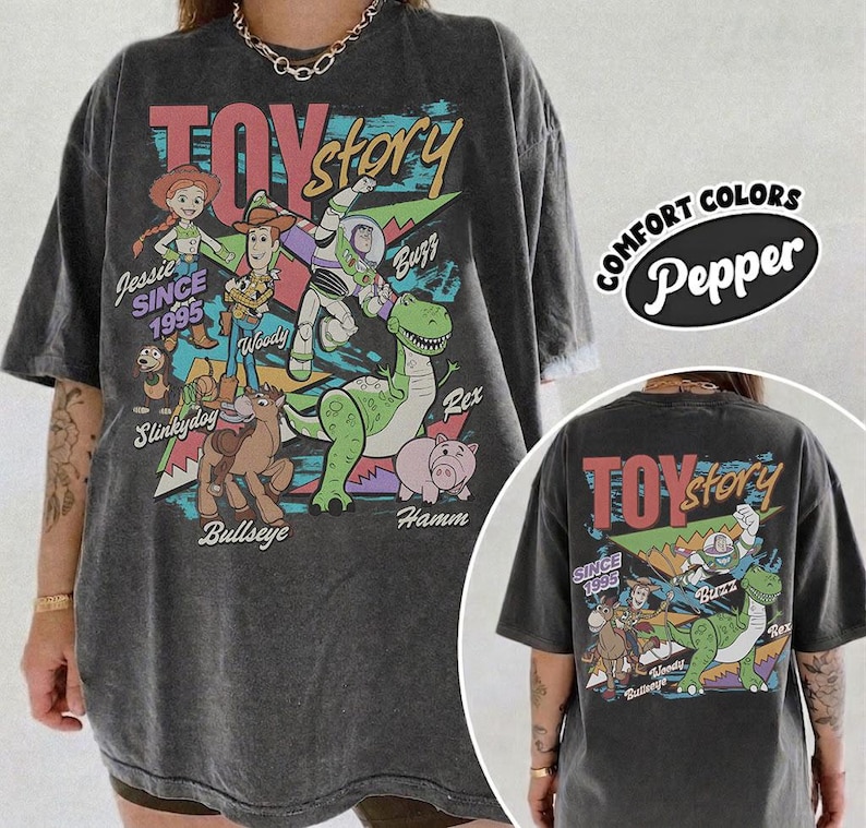 May include: A charcoal gray oversized t-shirt featuring a vintage-style Toy Story graphic print. The design includes characters Jessie, Woody, Buzz Lightyear, Slinky Dog, Bullseye, Rex, and Hamm with the text Toy Story and Since 1995.