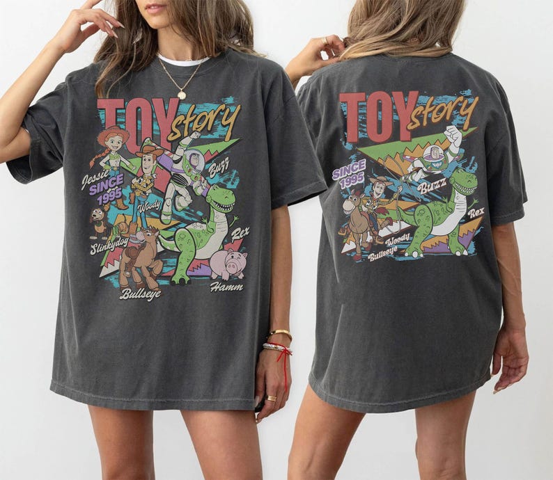 May include: A charcoal gray short-sleeve t-shirt featuring a graphic print of Toy Story characters including Woody, Buzz Lightyear, Jessie, Rex, Hamm, Bullseye, and Slinky Dog. The design includes the text Toy Story and Since 1995 in colorful lettering.