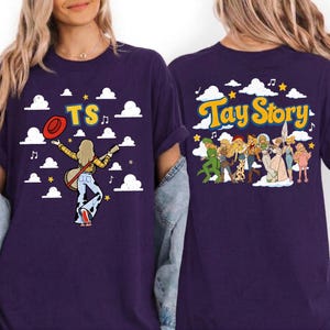 May include: A dark purple t-shirt featuring a graphic print on the front and back. The front shows a figure with a guitar, a red hat, and the text TS. The back displays a group of illustrated characters and the text Tay Story with clouds.