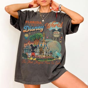 May include: A charcoal gray short-sleeve t-shirt featuring a colorful graphic print of Florida Disney World. The design includes Mickey Mouse, the castle, the Epcot sphere, and the text Florida Est. 1971 Disney World The Most Magical Place On Earth.