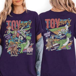 May include: A dark purple t-shirt featuring a graphic print of Toy Story characters including Jessie, Woody, Buzz Lightyear, Rex, Slinky Dog, Bullseye, and Hamm. The design includes the text Toy Story and Since 1995 with colorful geometric shapes.