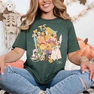 Puede incluir: Una camiseta de manga corta de color verde oscuro con un estampado gr&aacute;fico de varios personajes femeninos ilustrados con diversos atuendos. El texto TS aparece en amarillo sobre los personajes y una peque&ntilde;a bola colorida se ve abajo.