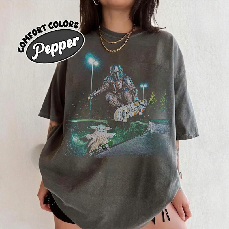 Op de afbeelding: Donkergrijs Comfort Colors T-shirt met een afbeelding van een sciencefictionpersonage dat aan het skateboarden is. De afbeelding bevat de tekst "Comfort Colors Pepper" in een witte ovaal. Het ontwerp toont een personage in harnas en een klein groen wezen.
