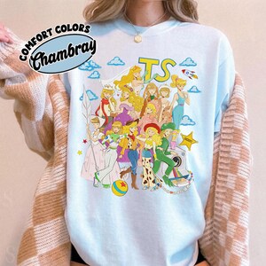 Puede incluir: Una sudadera de cuello redondo azul claro con un estampado gr&aacute;fico de varios personajes rubios con diversos atuendos, incluyendo un traje espacial, ropa de vaquera y vestidos. El texto "COMFORT COLORS" y "Chambray" aparece en un logotipo circular arriba a la izquierda.