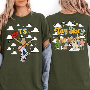 May include: An olive green t-shirt featuring a graphic print. The front displays a figure with a guitar, a red hat, and the text TS amidst white clouds. The back shows a group of illustrated figures and the text Tay Story with clouds.