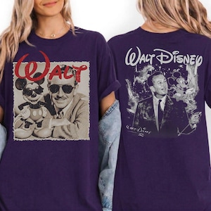 May include: A dark purple t-shirt featuring a graphic on the front of Walt Disney and Mickey Mouse wearing sunglasses with the red text WALT above them. The back of the shirt displays a black and white illustration of Walt Disney with the text WALT DISNEY.
