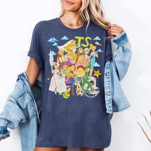 Puede incluir: Una camiseta jaspeada de color azul oscuro con un estampado gr&aacute;fico de varios personajes animados con diversos atuendos, que incluye un logotipo amarillo 'TS' en la parte superior, un cohete, nubes, una estrella amarilla y una c&aacute;mara blanca con rayas arco&iacute;ris.