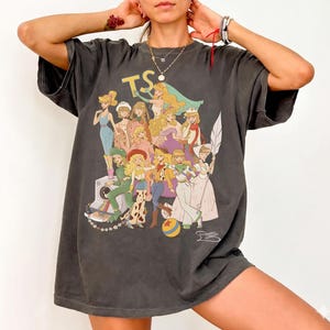 Puede incluir: Una camiseta de manga corta gris carb&oacute;n con un gran estampado gr&aacute;fico de varios personajes animados en estilo collage. El dise&ntilde;o incluye las letras TS en la parte superior y una pelota colorida en la parte inferior, con figuras en diversos atuendos.