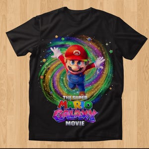 May include: A black short-sleeve t-shirt featuring a graphic of Mario with arms outstretched in front of a colorful swirling galaxy background. Below the character, the text reads THE SUPER MARIO GALAXY MOVIE in bold, multi-colored, block lettering.