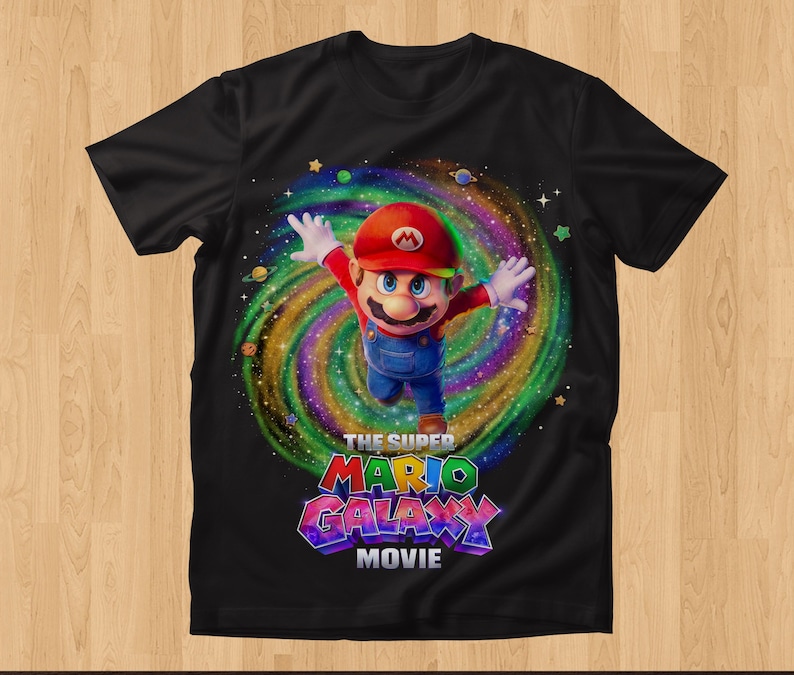 May include: A black short-sleeve t-shirt featuring a graphic of Mario with arms outstretched in front of a colorful swirling galaxy background. Below the character, the text reads THE SUPER MARIO GALAXY MOVIE in bold, multi-colored, block lettering.