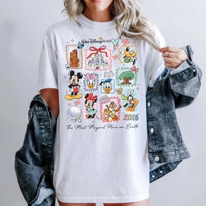 Może przedstawiać: Biała koszulka z kolorowymi postaciami Disneya oraz napisami "Walt Disney World" i "2026". Koszulka zawiera r&oacute;wnież frazę "The Most Magical Place on Earth".